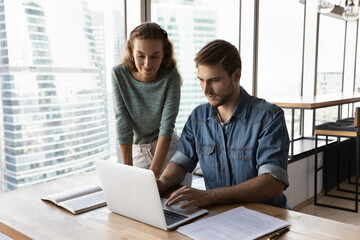 Friendly business mentor training intern, looking at laptop screen, supervising work process,...