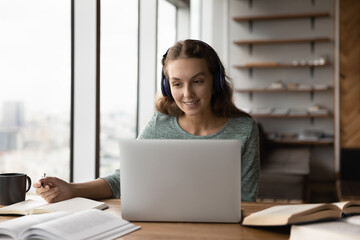 Happy gen Z student girl in headphones involved in online learning event, watching webinar on...