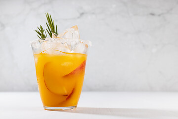Healthy summer juice - peach cocktail with ice, rosemary twig, sugar rim, fruit slices in misted glass in elegant white interior on soft light wood table.
