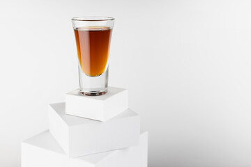 Golden brandy shot glass in hard light with shadow, reflection on high stack of white boxes, alcohol drink in rich elegant minimal geometric style.