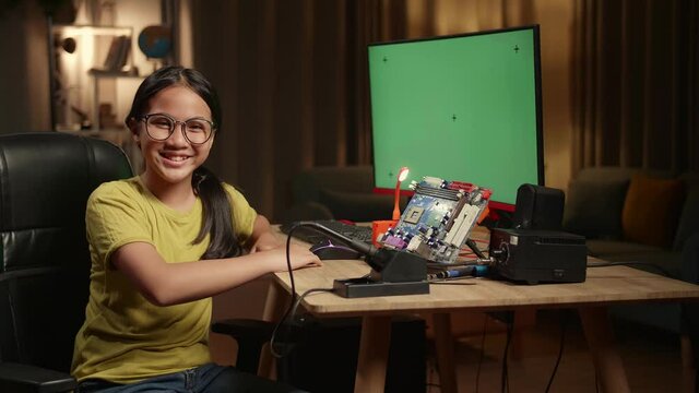 It Asian Girl Is Working With Desktop Computer And Mainboard In Home, Mock Up Green Screen Display. She Turns And Warmly Smiles Into The Camera, Genius Children Concept
