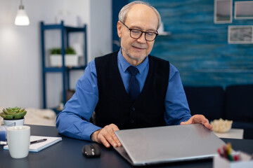 Businessman with gray hair opening laptop to start working. Elderly man entrepreneur in home workplace using portable computer sitting at desk while wife is holding tv remote.