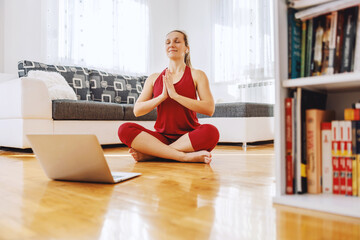 Calm yoga instructor sitting in lotus yoga pose on the floor at home and having an online class.