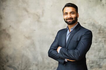 Smiling middle-age Indian high skilled leader in formal suit looks at the camera, confident ambitious bearded arabic businessman stands in office with arms folded on the grey background - obrazy, fototapety, plakaty