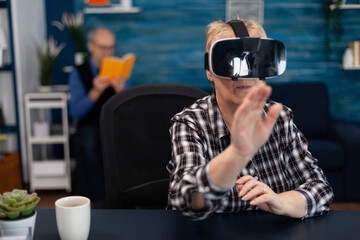 Happy senior woman wearing virtual reality headset doing hand gesture. Cheerful elderly woman experiencing augmented reality cyberspace while husband is reading a book on sofa.