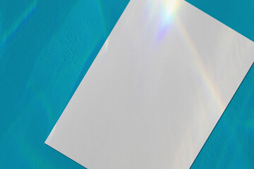 Close up of empty white vertical rectangle a4 poster or business card mockup, lying diagonally with overlay of rainbow light refraction caustic effect and shadow on trendy blue concrete background