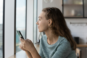 Thoughtful gen Z teen girl holding smartphone, looking out window in deep thoughts, thinking over...