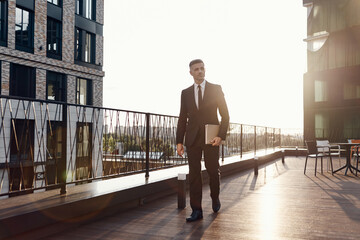 Fototapeta na wymiar Confident mature man in full suit carrying laptop while walking by business center terrace outdoors