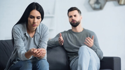 Fototapeta na wymiar worried woman sitting and looking at hands near blurred and displeased boyfriend quarrelling in living room