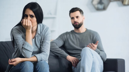 worried woman covering face and sitting near blurred and displeased boyfriend quarrelling in living room