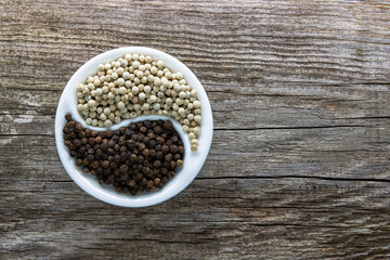 A porcelain bowl, divided in half, filled with black and white peppercorns on an aged rough board