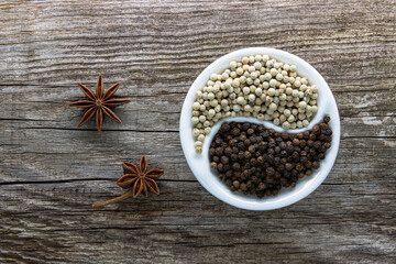 A halved porcelain bowl filled with black and white peppercorns on an aged rough board. Star anise...