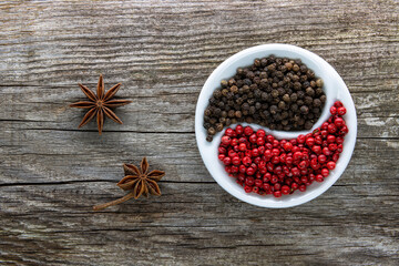 A halved porcelain bowl filled with black and red peppercorns on an aged rough board. Star anise inflorescences lie nearby
