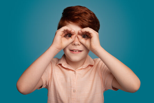 Ginger boy gesturing binocle sign on studio in trendy style. Fashion model.