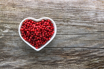 A heart-shaped porcelain bowl filled with red peppercorns on an aged rough board.