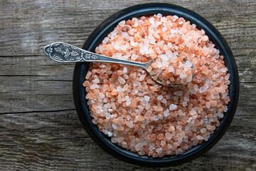 A round earthenware black bowl filled with large crystals of pink himalayan salt on an aged rough board. On top is a vintage silver spoon with salt