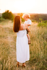 Beautiful woman in white light dress while walking with her son along a picturesque field at sunset