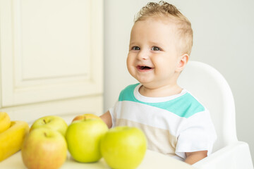 Fototapeta premium cute baby boy sitting at the table in child chair eating apple on white kitchen.
