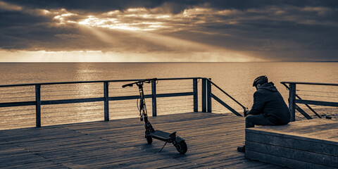 A man sits on the boardwalk next to his scooter watching the dramatic evening storm clouds at sunset