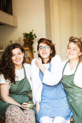 Three laughing young women in aprons in a pottery studio looking at the camera, close-up, daylight, vertical photo