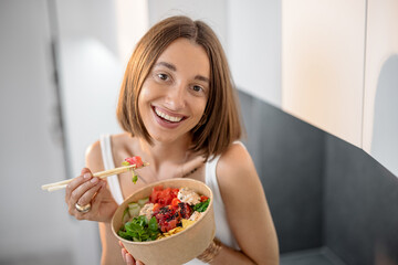 Portrait of a happy woman eating healthy asian bowl with tuna and salad on the modern kitchen at home. Healthy takeaway asian food and modern lifestyle at home
