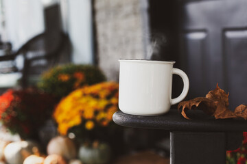 Steaming coffee cup sitting on arm of rocking chair on a front porch that has been decorated for autumn with heirloom pumpkins and mums. Selective focus with blurred background.