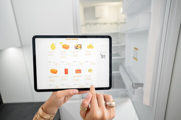Woman shopping groceries online using digital tablet while standing near the empty fridge on the kitchen. E-commerce, smart kitchen technology concept