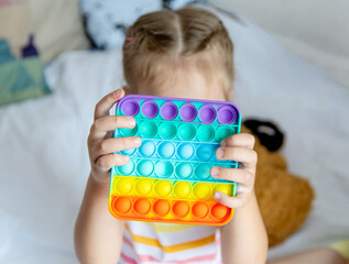 The girl holds in her hands in front of her face a rainbow toy anti-stress pop it.