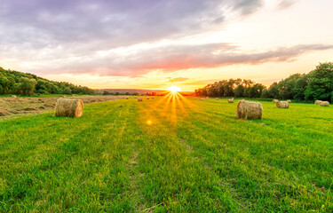 Scenic view at sunset or sunrise in green shiny field with hay stacks, bright cloudy sky, golden sun rays, summer valley landscape
