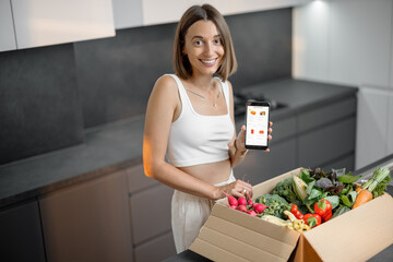 Woman buying fresh vegetables online, standing with package full of food and holding smart phone...