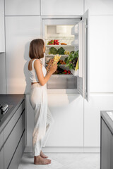 Young woman standing near the fridge full of fresh vegetables at modern kitchen. Healthy vegan...