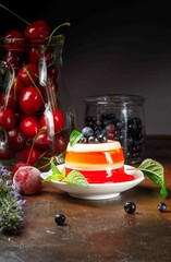 Berry jelly on a white plate, decorated with mint leaves. A glass jug with cherries and a glass with blueberries in the background.