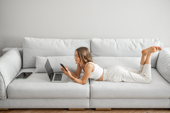 Young woman lying relaxed with phone and laptop on the comfortable couch at home. Front view. Working or leisure time at home