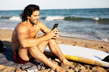 Handsome surfer with his surfboard. Young man using the phone while relaxing on the beach
