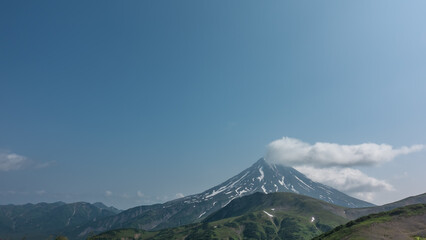 Fototapeta premium The conical stratovolcano Vilyuchinsky rises against the background of the blue sky. There is snow on the slopes. There is a fluffy white cloud over the top. A summer day. Kamchatka
