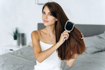 Healthy brunette woman brushing her hair with comb before the walk, while sitting at the bedroom and looking at the camera. Self care procedures concept