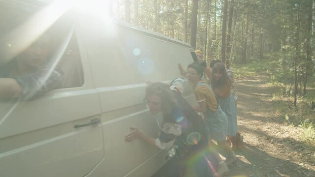 Company of young friends pushing van in forest, then clapping hands and embracing as car start riding