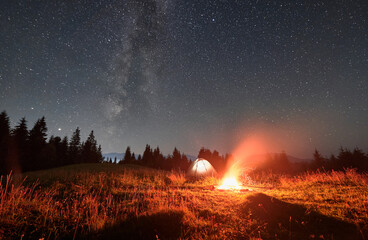 Beautiful landscape in the mountains. Tourist illulminated tent and campfire under night starry sky with Milky way. Mountains range behind spruces. Concept of night camping and astrophotography