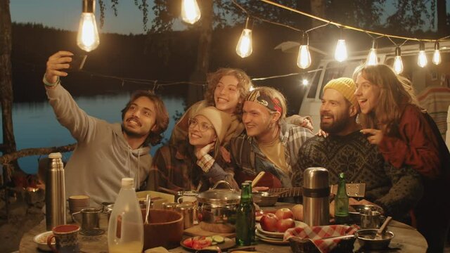 Company of young multiethnic friends smiling and posing for selfie at dinner table, then checking picture on smartphone while spending evening at campsite decorated with garland
