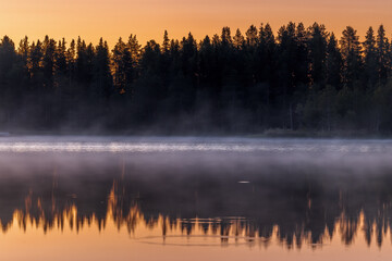 Colorful misty Sunset over lake with black silhouette of boreal forest, beautiful reflection, Kitkajärvi, Posio, Finland