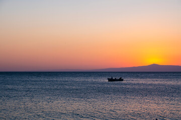 Fishermen in a tiny fisher boat with floating on peaceful blue Aegean Sea during sunrise, sunset. Dramatic orange sky and romantic island background with large copy space. Tranquility and serenity.
