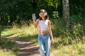A teenager walks through the woods and listens to music with headphones. A girl is walking along the path against the background of green trees.