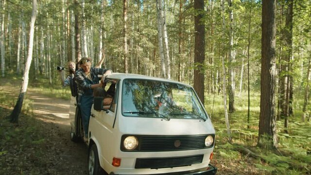 Company of young excited friends standing in van with opened door, smiling and taking pictures during ride on forest road