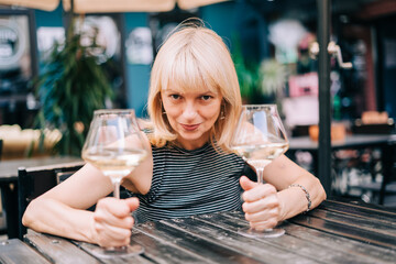 Happy funny adult mature woman sitting in bar outdoors and holding wine glasses in blurry restaurant background scene, drinking white wine. Summer sunny day on patio. People lifestyle