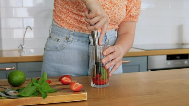 drinks and people concept - happy smiling young woman making strawberry mojito cocktail at home kitchen
