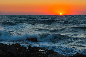 Sunset and storm at sea. Big waves against the setting sun. A summer storm in the ocean. Beautiful sea spray with foam breaks on the rocky shore. Beautiful natural background. The orange setting sun.