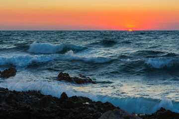 Fototapeta premium Sunset and storm at sea. Big waves against the setting sun. A summer storm in the ocean. Beautiful sea spray with foam breaks on the rocky shore. Beautiful natural background. The orange setting sun.