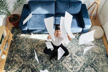 Education, graduation and people concept - top view of happy male student throwing a bunch of sheets at home showing his emotions