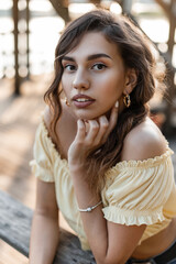 Summer portrait of a young beautiful woman model with curly hairstyle fashion yellow top blouse stands on pier near a lake