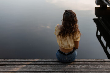 Pretty young woman in jeans and a blouse sits and rest on a wooden pier near the lake